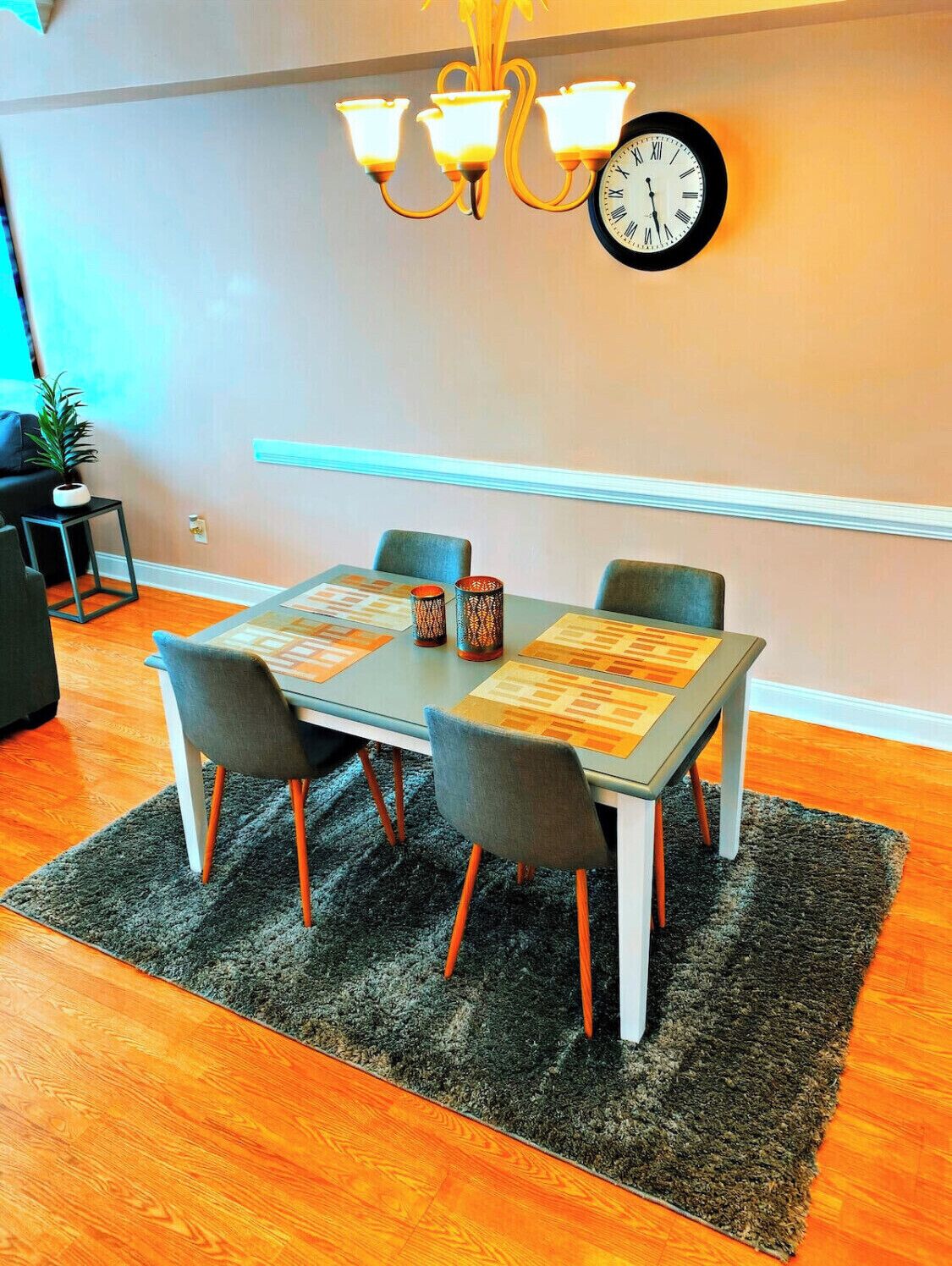 A dining area with a table set for four, a wall clock, and a chandelier.