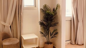 Una habitación con una silla color beige, un estante pequeño blanco, una planta en maceta y una ventana con cortinas color beige.
