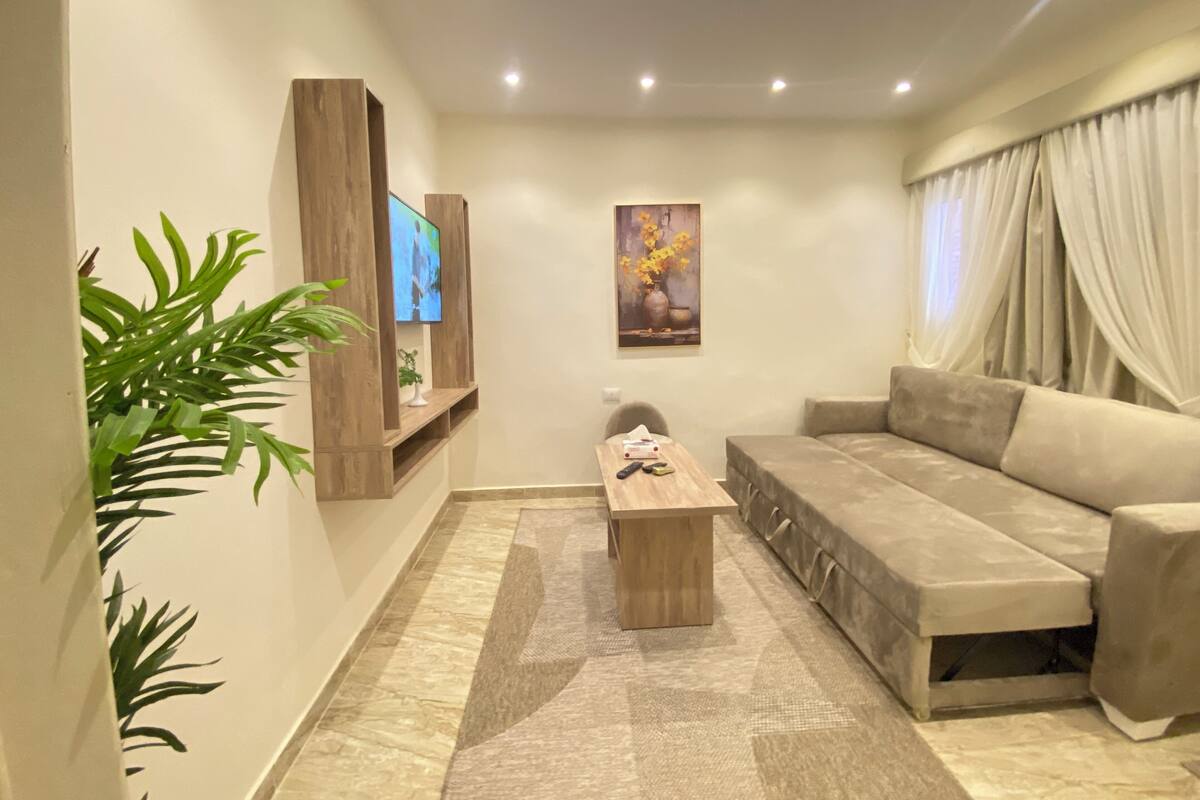 A modern living room with a sofa, a coffee table, a TV mounted on a wooden panel, a painting on the wall, and a large plant.