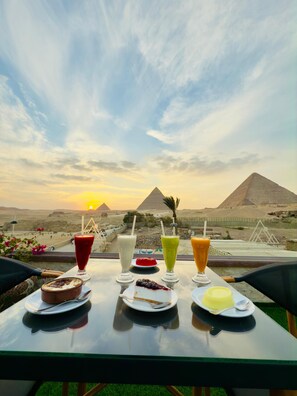 Une table dressée avec diverses boissons et desserts, le tout mis en valeur par un décor composé des pyramides et d’un coucher de soleil.