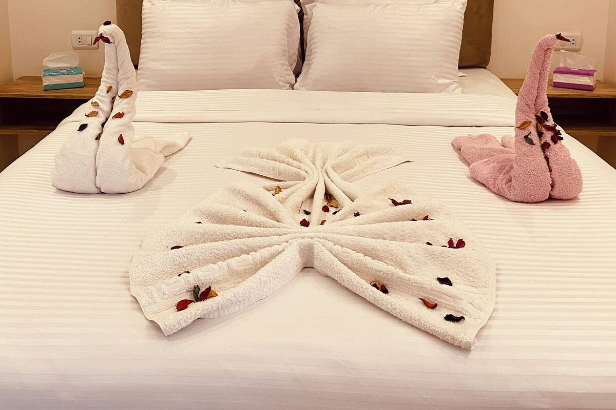 A hotel room with a bed, pillows, a painting of a lake, and two towels shaped like swans with flowers.