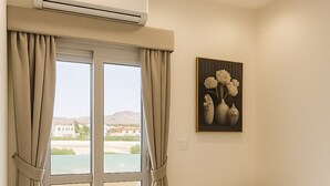 Una habitación con puerta corrediza de vidrio, cortinas color beige, un pouf redondo, una mesita auxiliar blanca y una obra enmarcada en la pared.