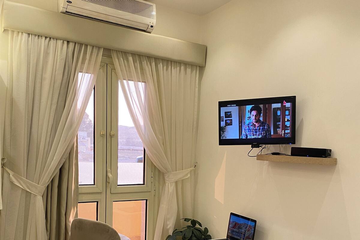 A hotel room with a bed, a desk with a laptop, a TV on the wall, and a large window with curtains.