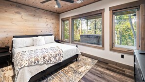 Une chambre à coucher avec un grand lit, un plafond en bois et deux fenêtres donnant sur la forêt.