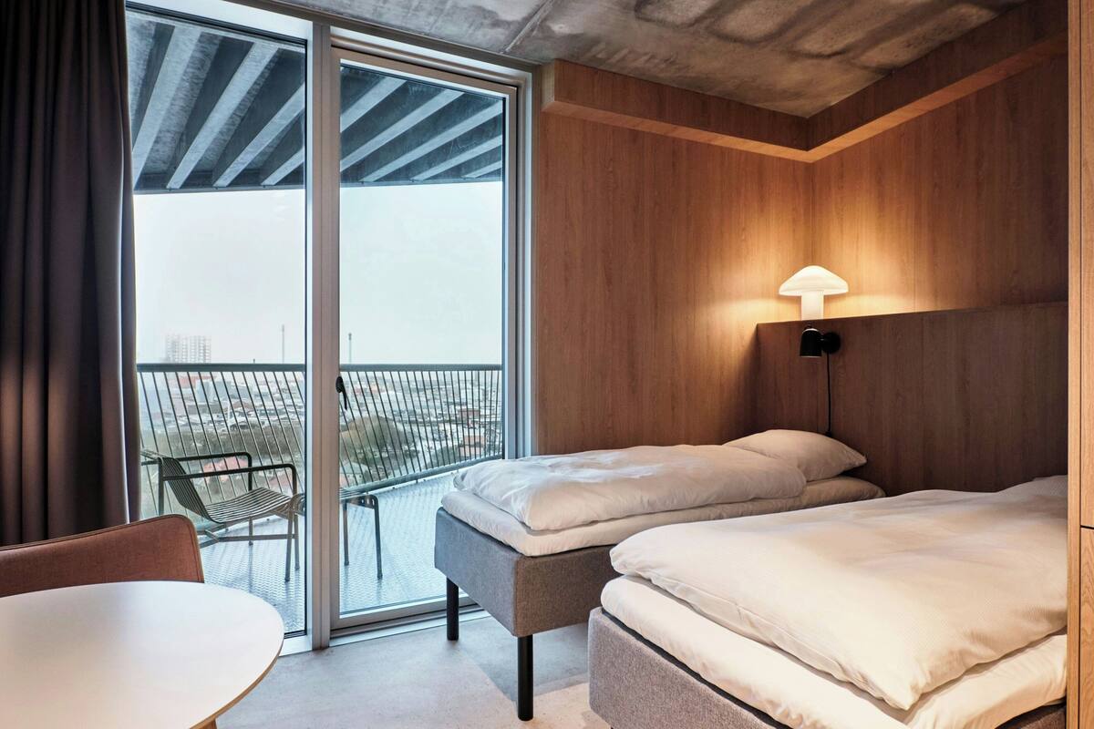 Une chambre d’hôtel moderne dotée d’une grande porte-fenêtre coulissante donnant sur un balcon extérieur, d’un lit avec une tête de lit, d’une table de chevet et d’une lampe.