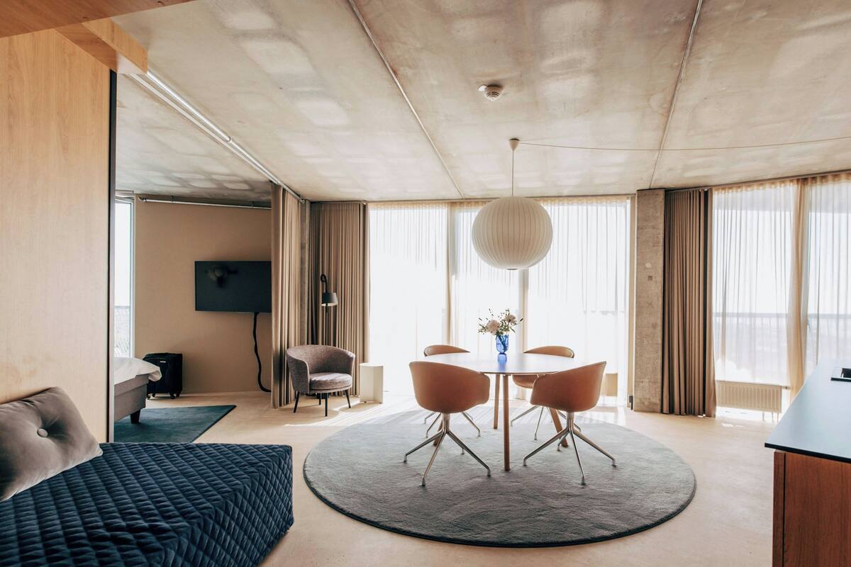 Une chambre d’hôtel moderne avec une table à manger ronde, deux chaises, un canapé, un téléviseur et une grande fenêtre avec des rideaux.