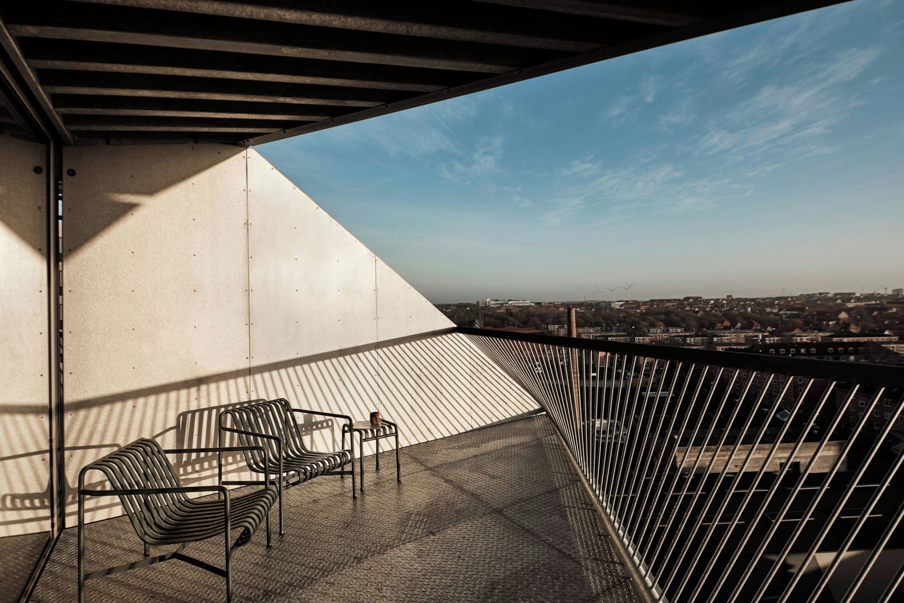 Un balcon avec des chaises et une table en métal, d’où l’on peut admirer un paysage urbain avec des immeubles et des éoliennes.