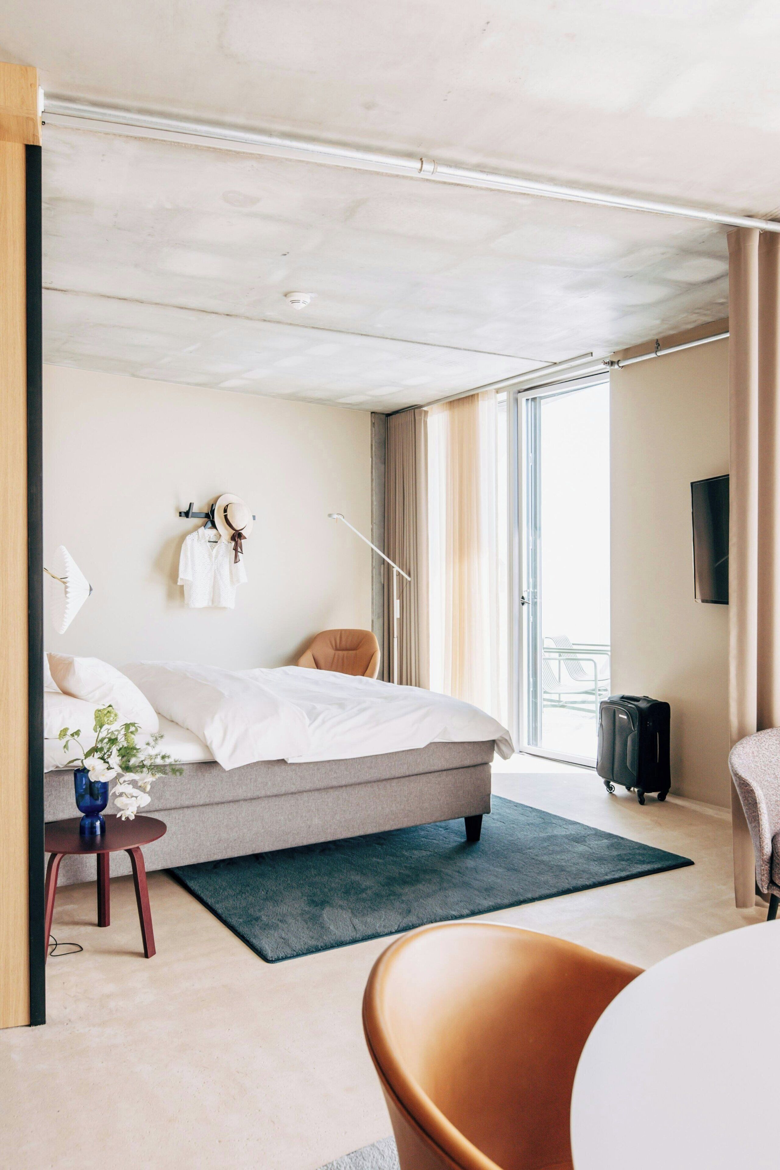 Una habitación de hotel moderna con una cama, un escritorio, una silla, un televisor y un ventanal amplio.