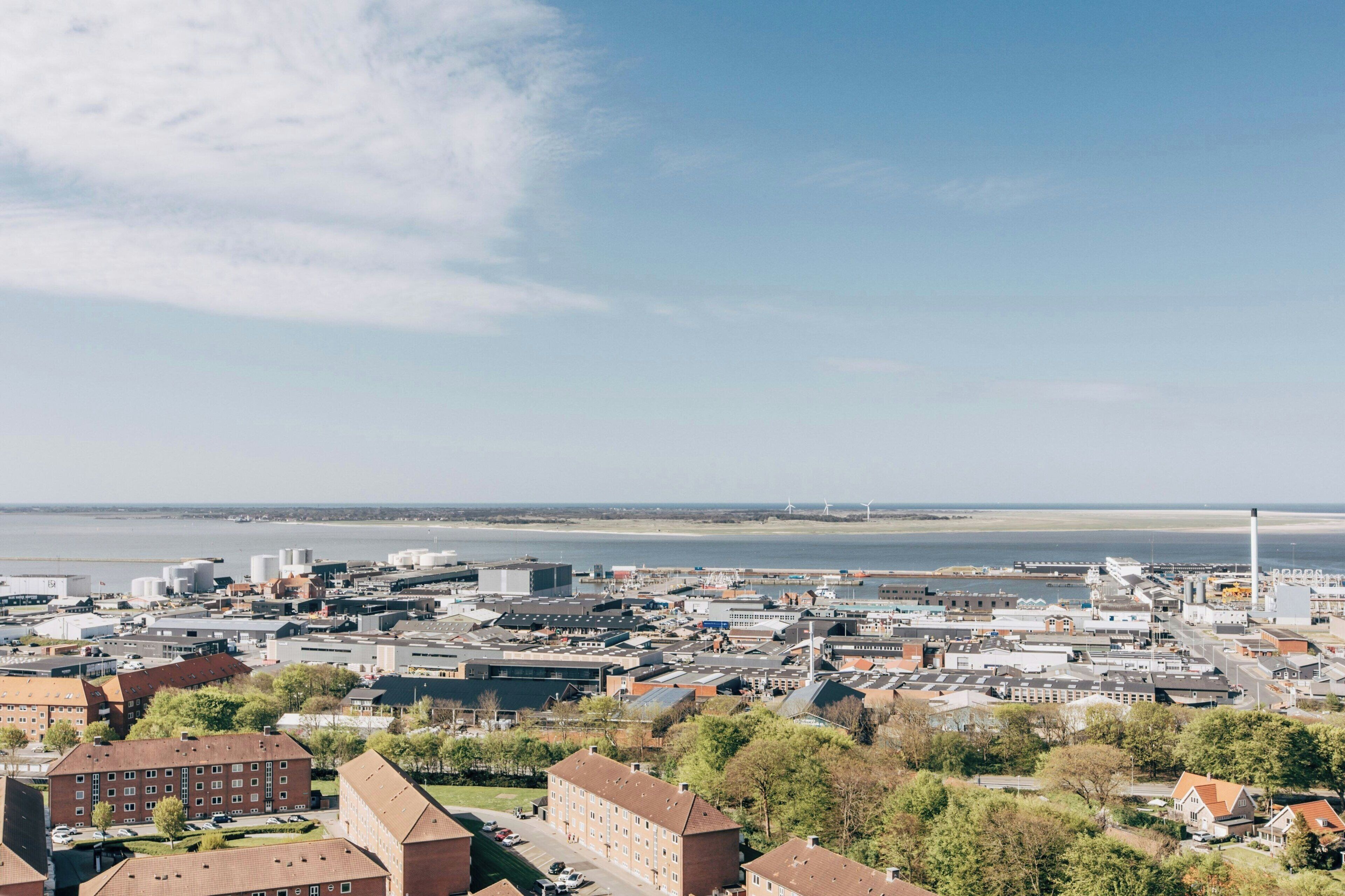 Un paisaje urbano costero con edificios industriales, una extensa masa de agua y un faro destacado.
