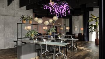 Ein modernes Restaurant mit hoher Decke, freiliegenden Balken und einer Neonlicht-Installation. Tische und Stühle sind großzügig angeordnet, mit Topfpflanzen und dekorativen Laternen.