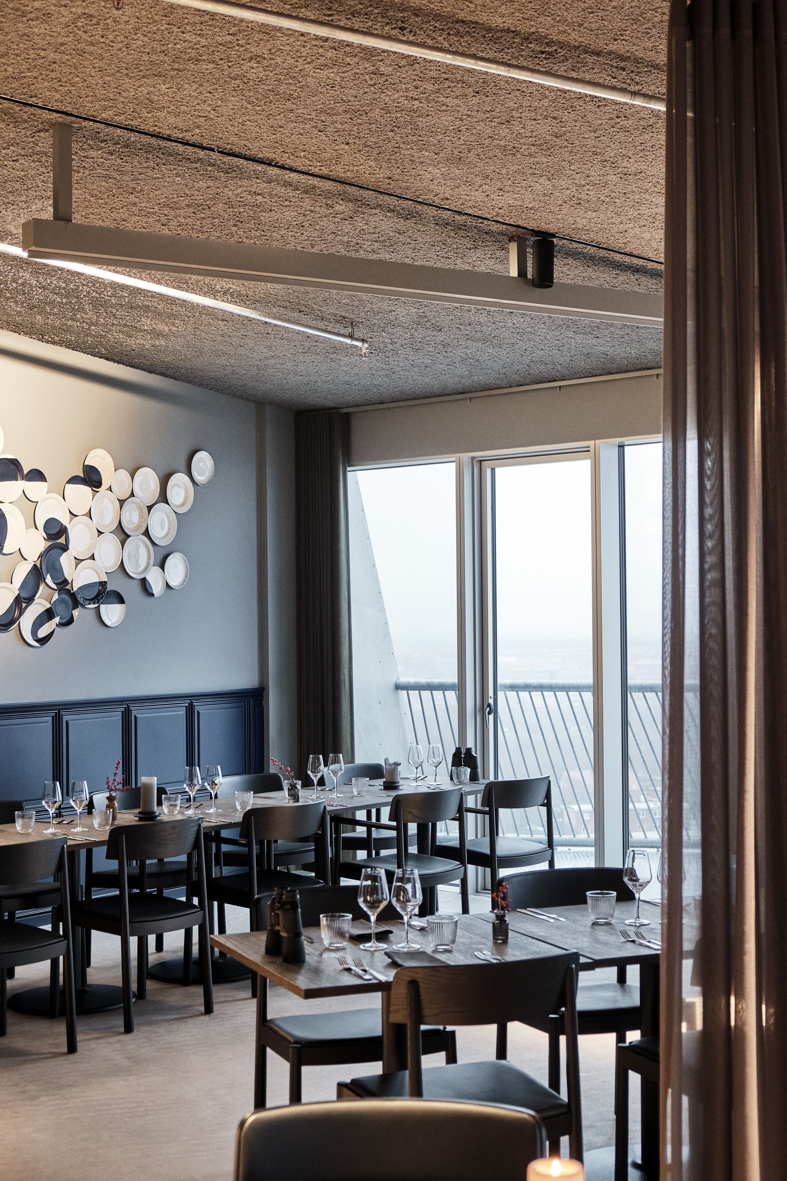 A modern dining area with multiple tables and chairs, a decorative wall with plates, and large windows offering a view.