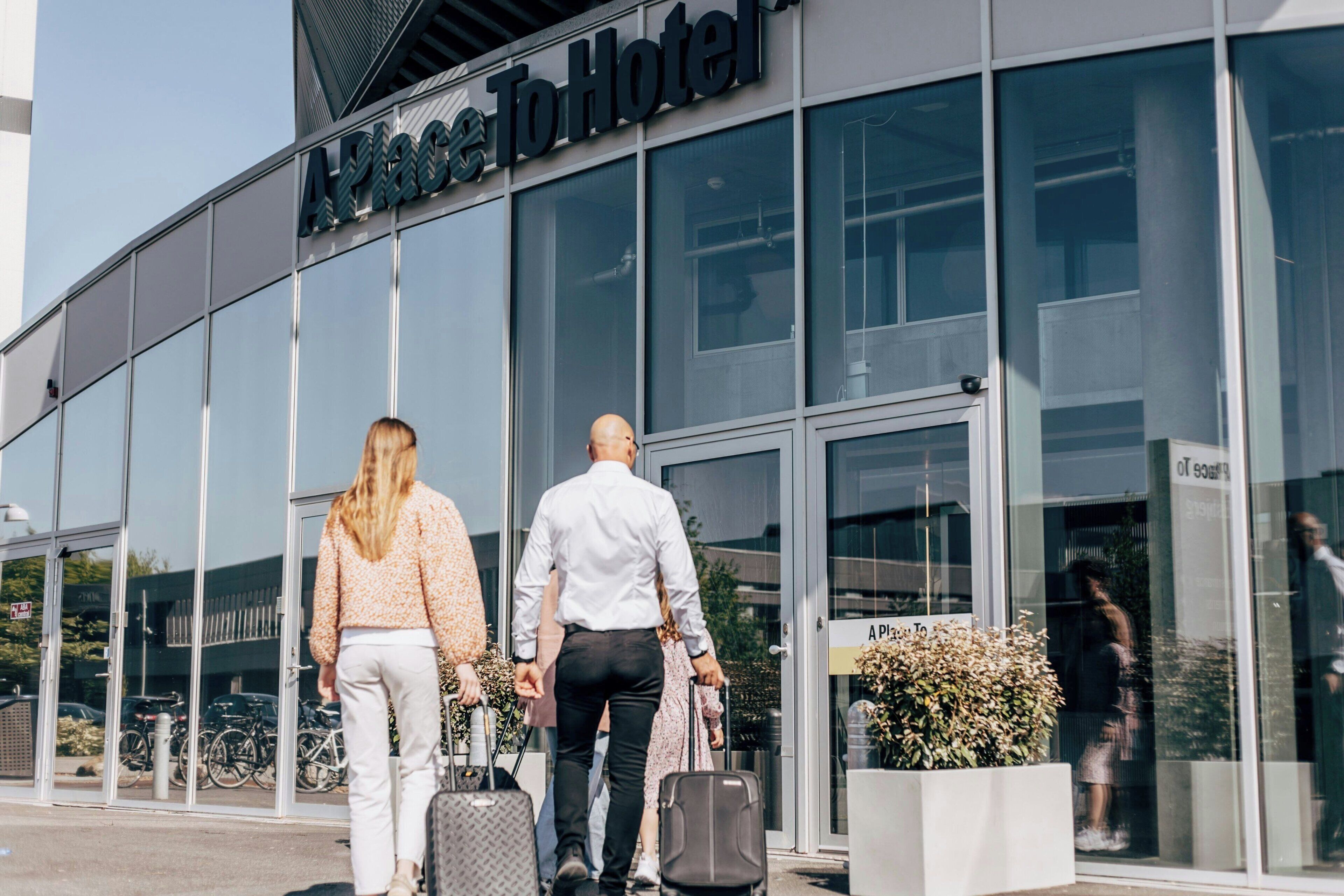 A modern hotel entrance with glass doors, a sign reading 'APAC To Hotel,' and a few people walking in and out.
