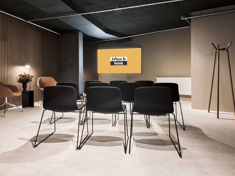 A conference room with a projection screen displaying "A Place To Hotels," black chairs arranged around a table, and a wooden slatted wall with a vase of flowers.