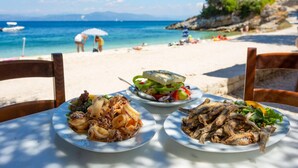 Une table dressée avec des assiettes de nourriture, une plage avec des personnes à l’arrière-plan, et une vue dégagée sur la mer et les montagnes.