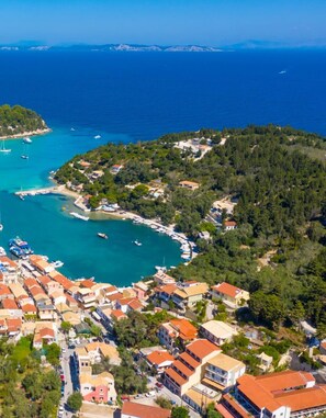 An aerial view of a coastal town with a marina, houses with red roofs, and a hilly backdrop.
