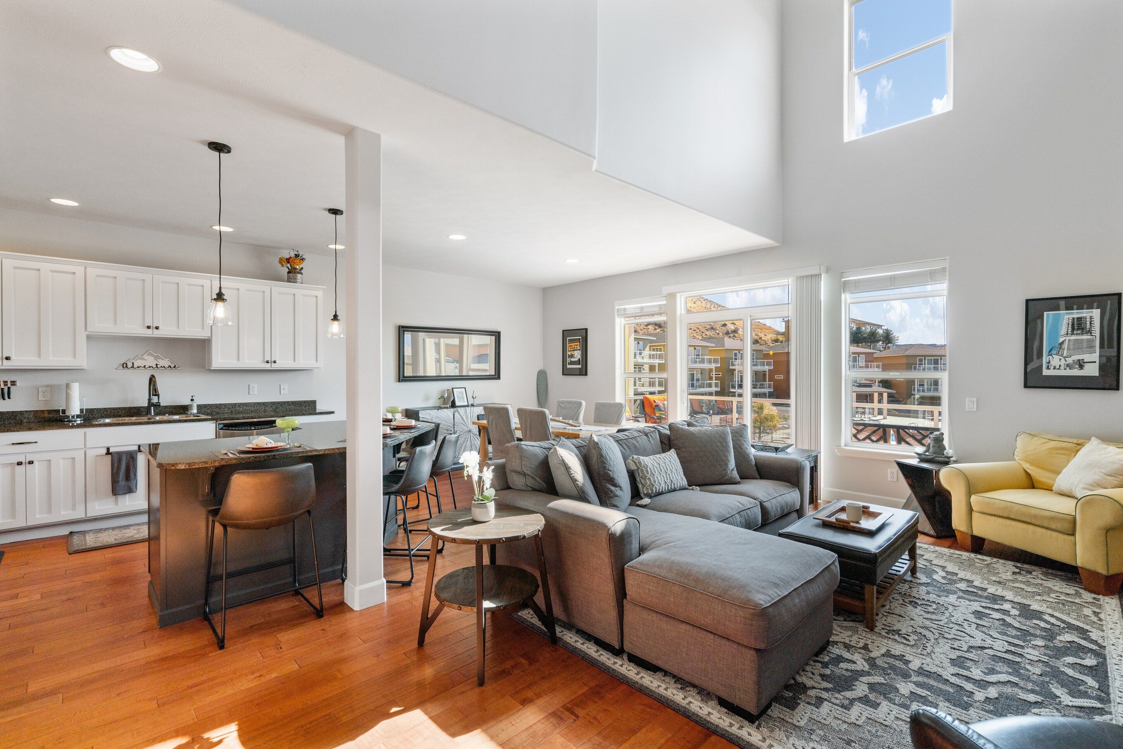 A spacious living area with a grey sectional sofa, a yellow armchair, and a kitchen with white cabinets and a bar area.