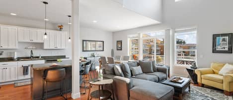 A spacious living area with a grey sectional sofa, a yellow armchair, and a kitchen with white cabinets and a bar area.