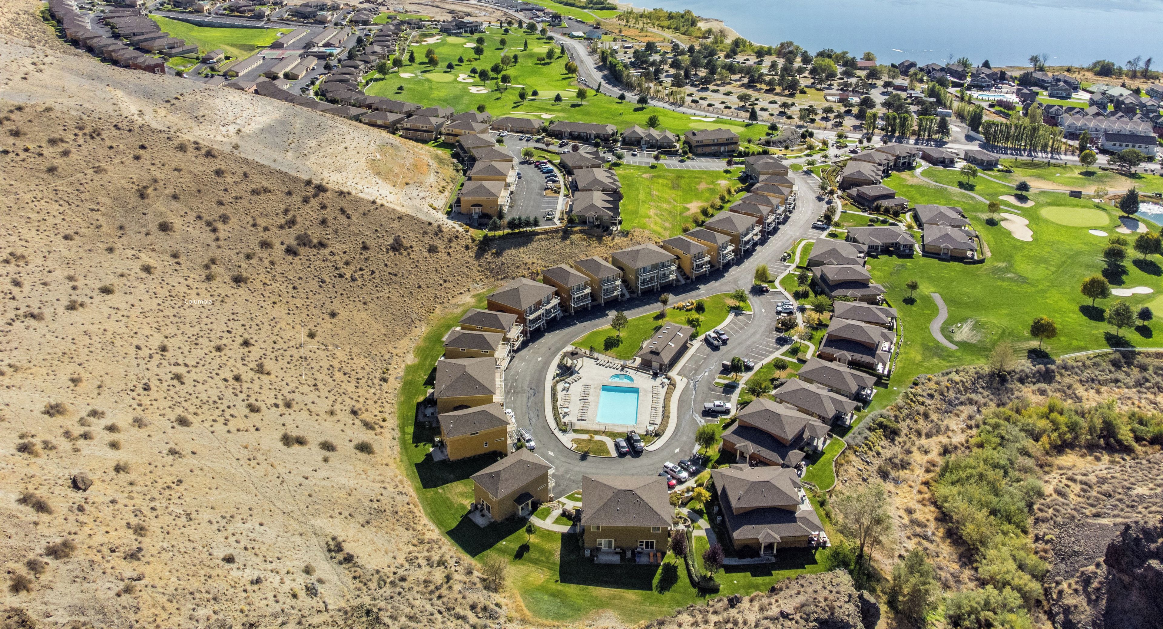 An aerial view of a residential area with houses arranged in a semi-circular pattern around a central swimming pool, with the Columbia River visible in the background.