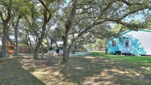 A backyard with large trees, a blue house, and a covered area.
