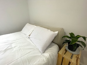 Uma cama bem arrumada com roupa de cama branca, uma planta em vaso sobre um banquinho de madeira e uma parede lisa.