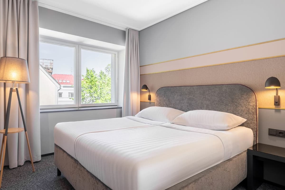 A hotel room with a bed, a window with curtains, a floor lamp, and a wall-mounted lamp.