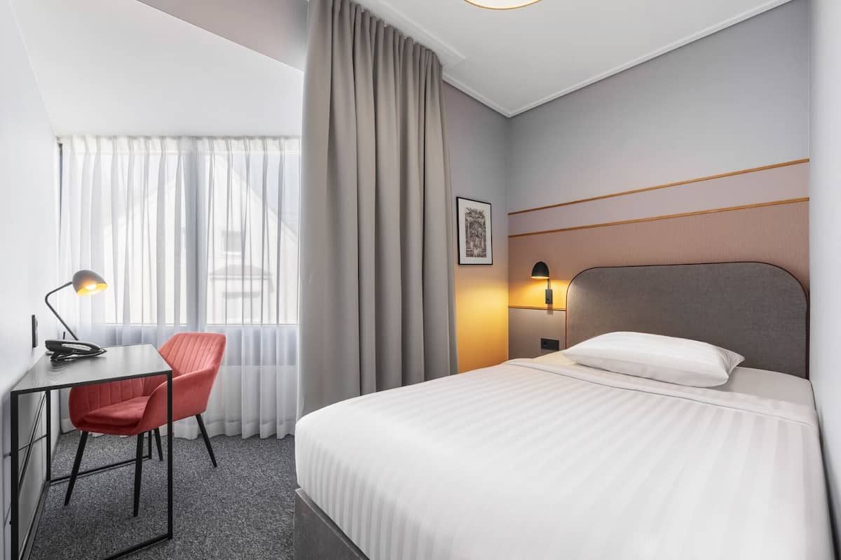 A hotel room with a bed, a desk with a chair, a lamp, and a window with curtains.