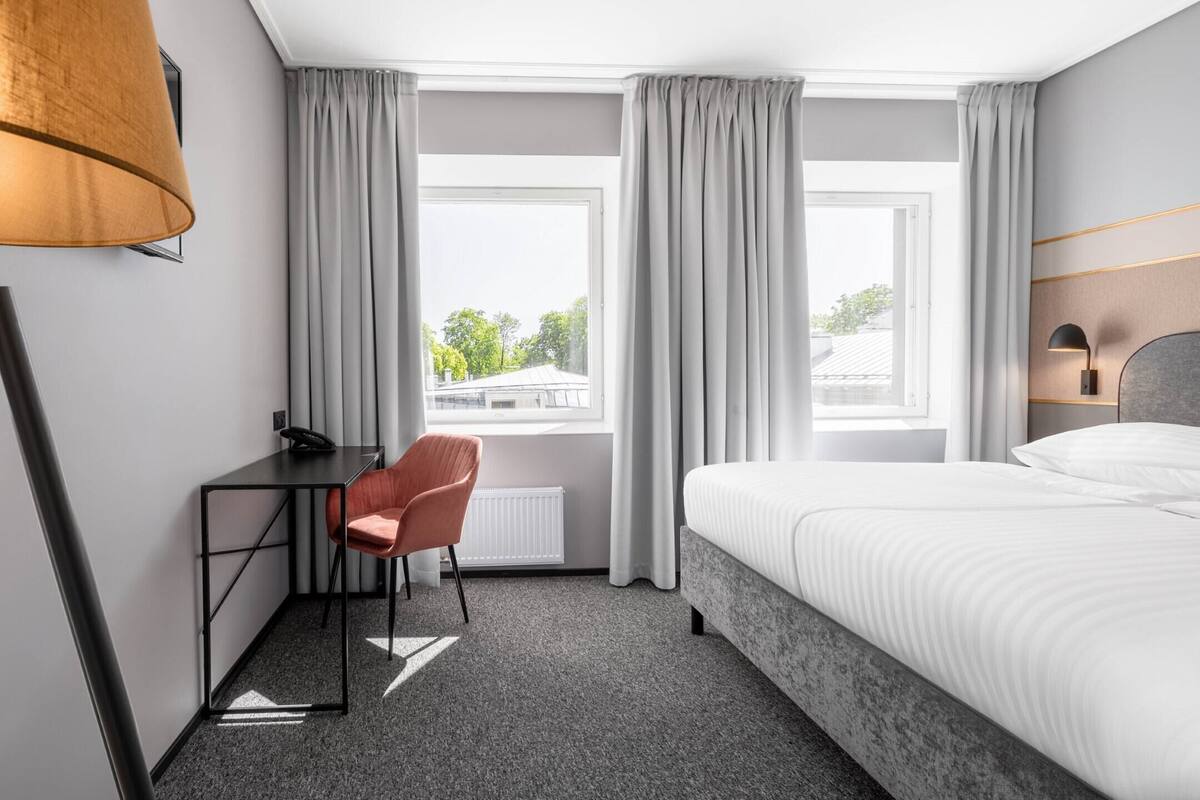A hotel room with a bed, a desk with a chair, a lamp, and a window with curtains.