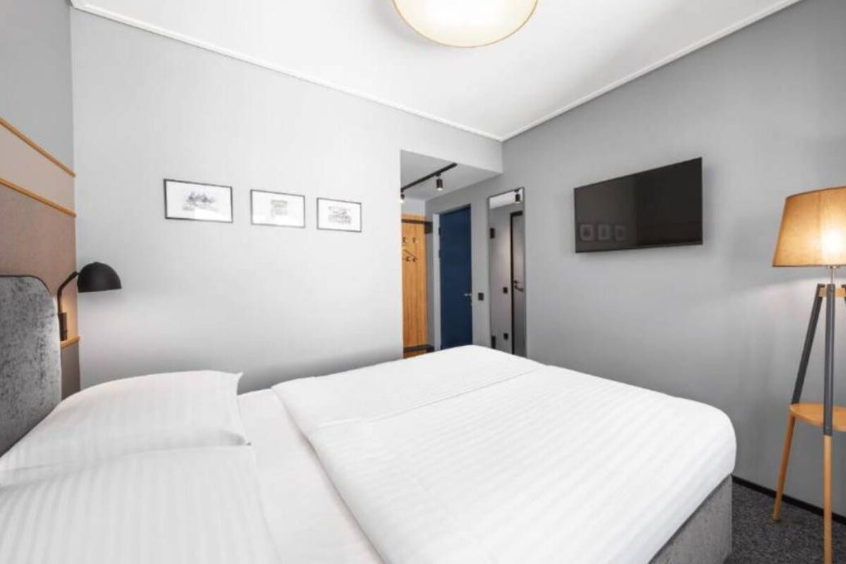 A hotel room with a bed, a desk, a chair, a lamp, and a television on the wall.