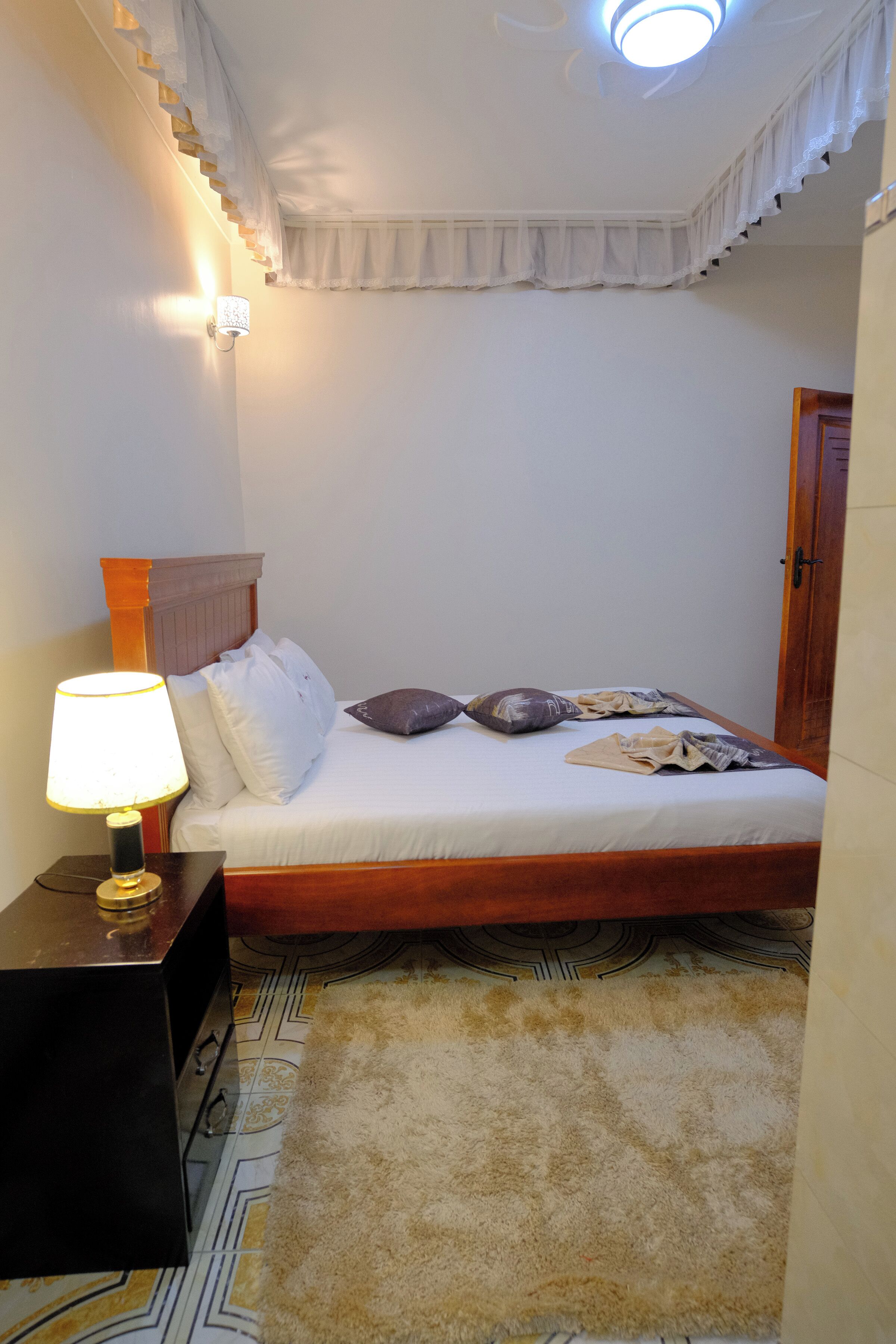 A bedroom with a bed, a nightstand with a lamp, a patterned rug, and a ceiling light.