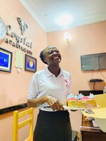 Una persona sosteniendo un pastel en una habitación con pared color melocotón, un reloj y diplomas enmarcados.