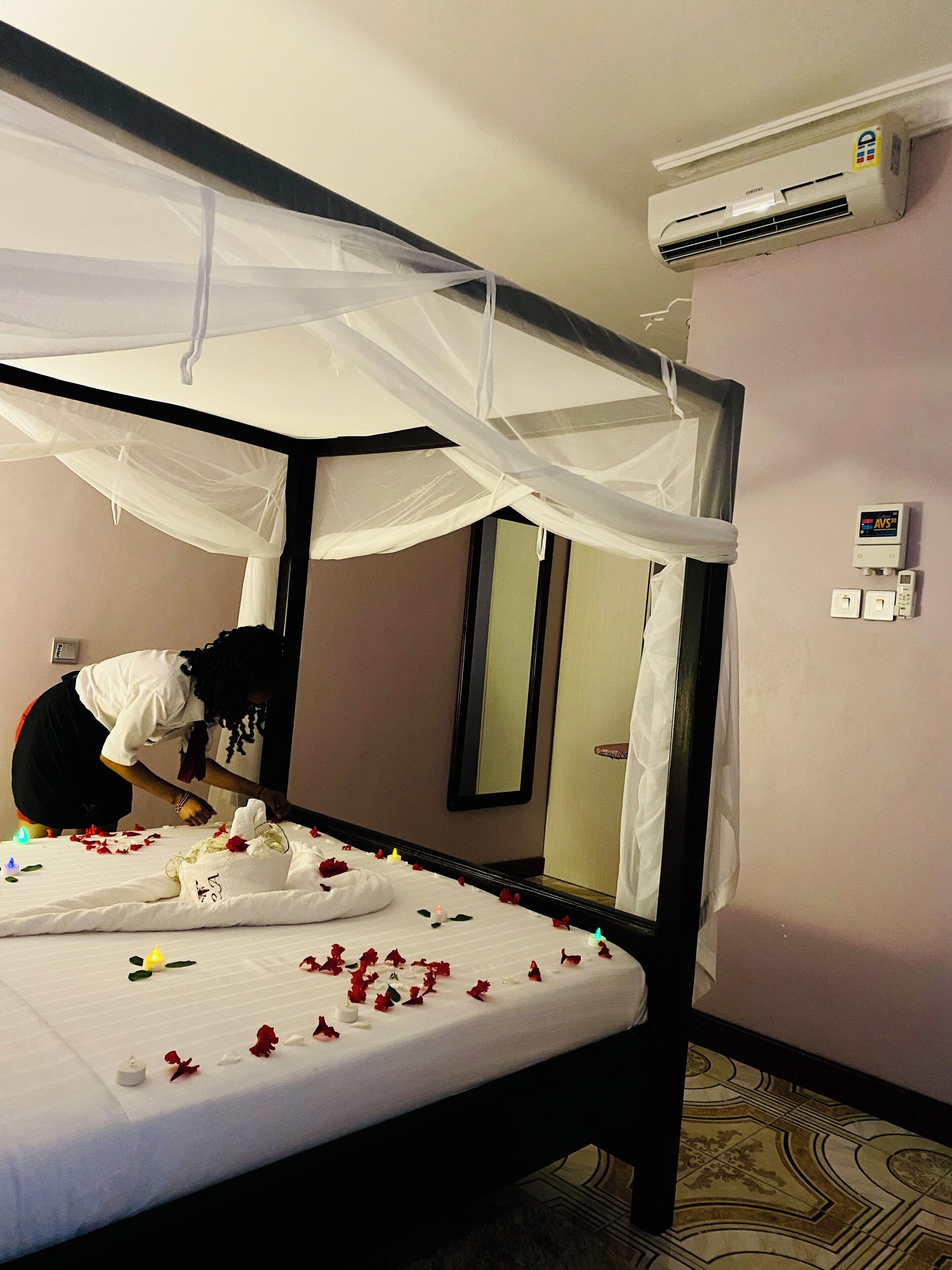 A hotel room with a four-poster bed, an air conditioning unit, a wall-mounted control panel, and a mirror.