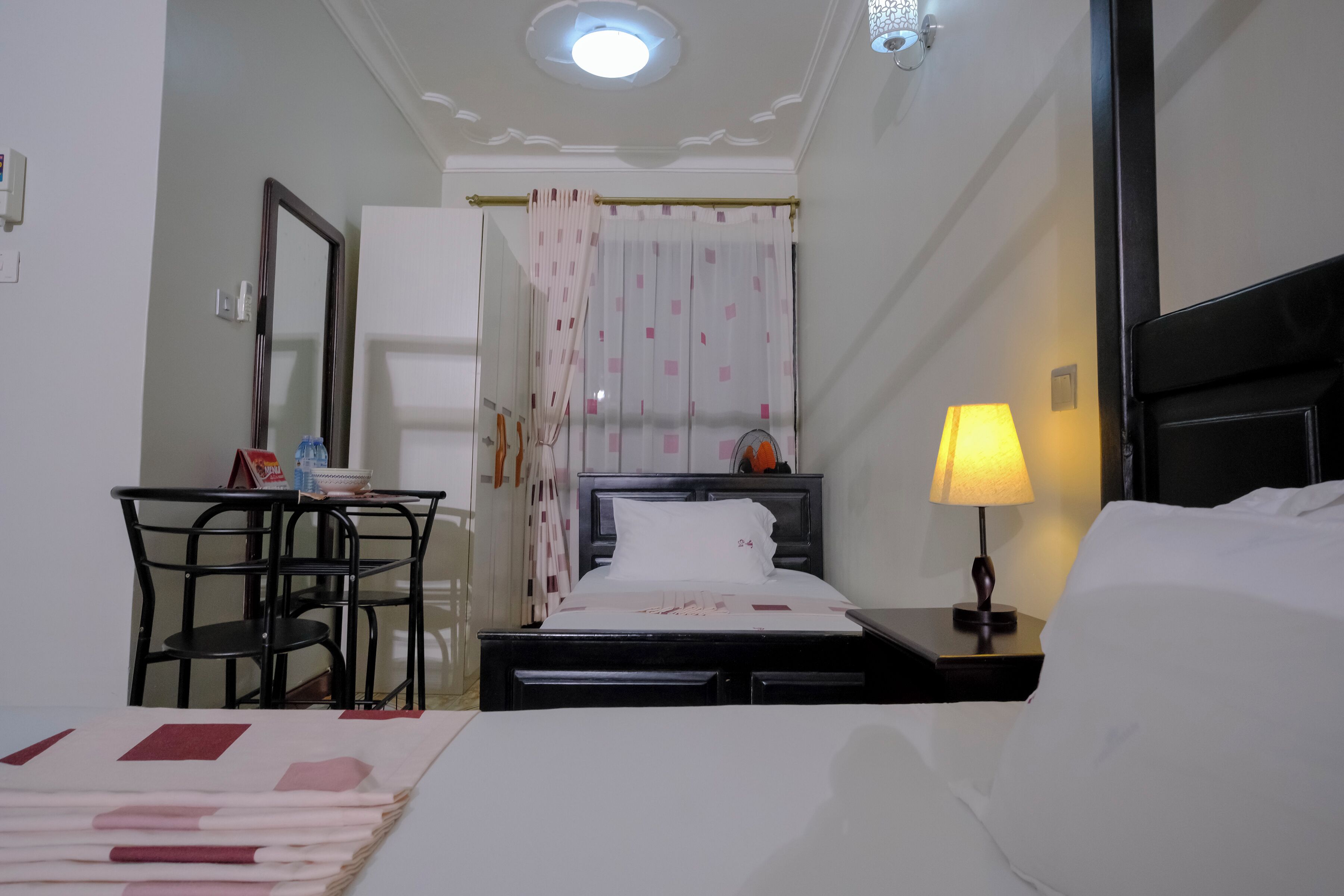 A hotel room with a bed, a desk, a chair, a nightstand with a lamp, and a window with curtains.