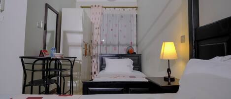 A hotel room with a bed, a desk, a chair, a nightstand with a lamp, and a window with curtains.