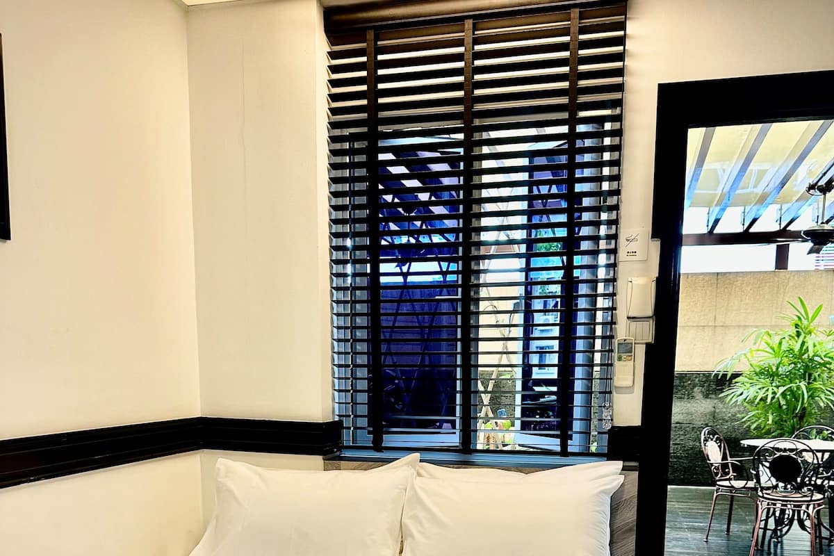 Ein Hotelzimmer mit einem großen Bett, einem Fenster mit Jalousien und Blick auf einen Außenbereich mit Stühlen und einem Tisch.