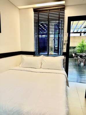 Une chambre d’hôtel avec un grand lit, une fenêtre avec des stores et une vue sur un espace extérieur aménagé avec des chaises et une table.