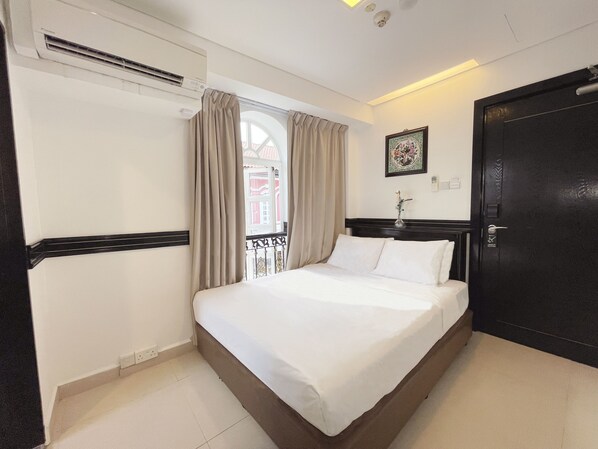 Une chambre d’hôtel comprenant un lit, une fenêtre avec des rideaux, un appareil de climatisation, une porte et un tableau accroché au mur.