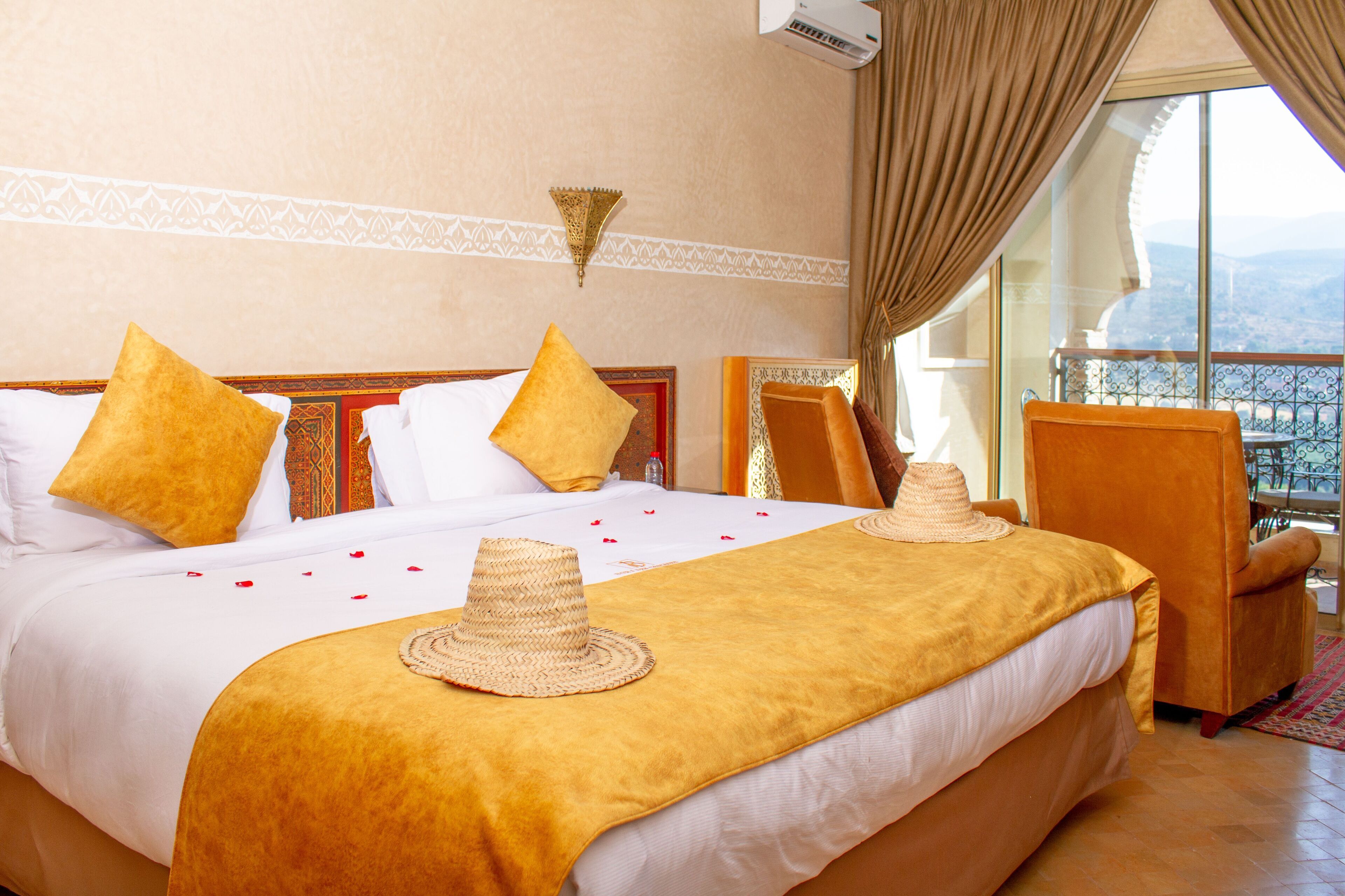 A hotel room with a large bed, two chairs, a balcony with a view, and a decorative wall lamp.