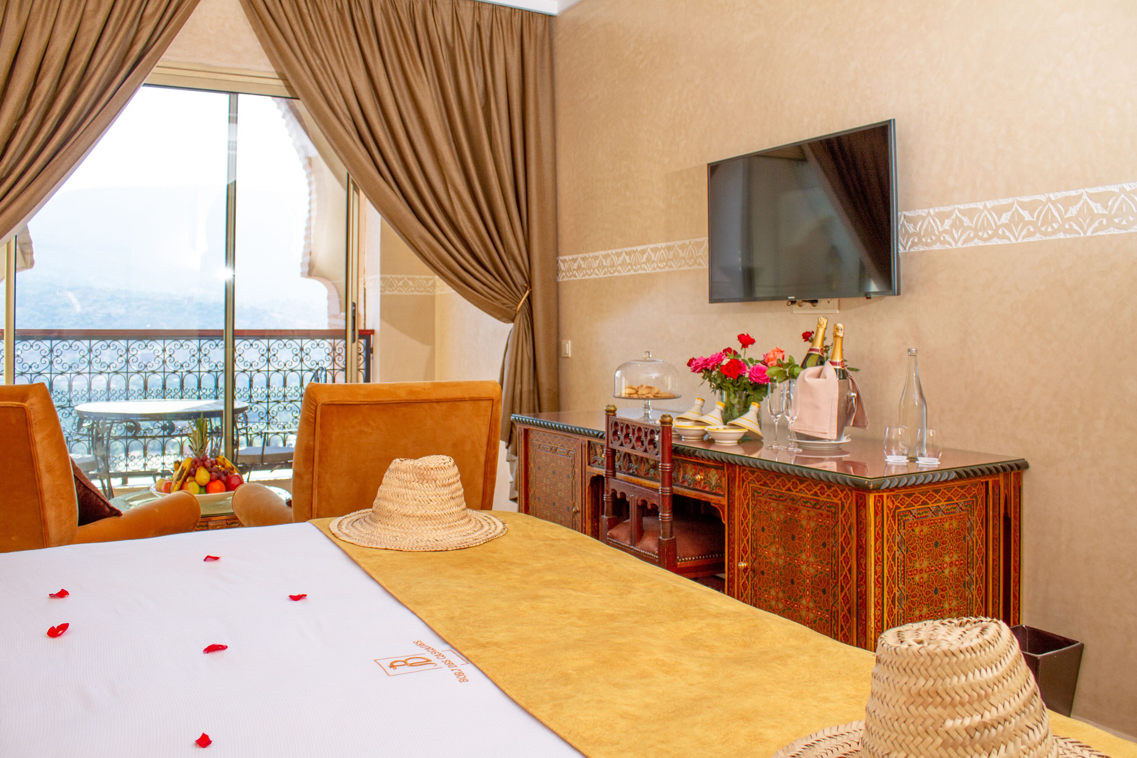 A hotel room with a balcony, a dining table with a white tablecloth, a television mounted on the wall, and a decorative cabinet with a mirror.