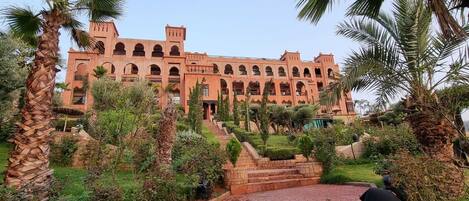 A large, multi-story building with arched windows and a central tower, surrounded by well-maintained gardens and palm trees.