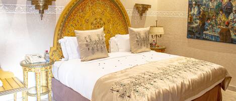 A large bed with ornate headboard, multiple pillows, a bedside table with a lamp, and a painting on the wall.