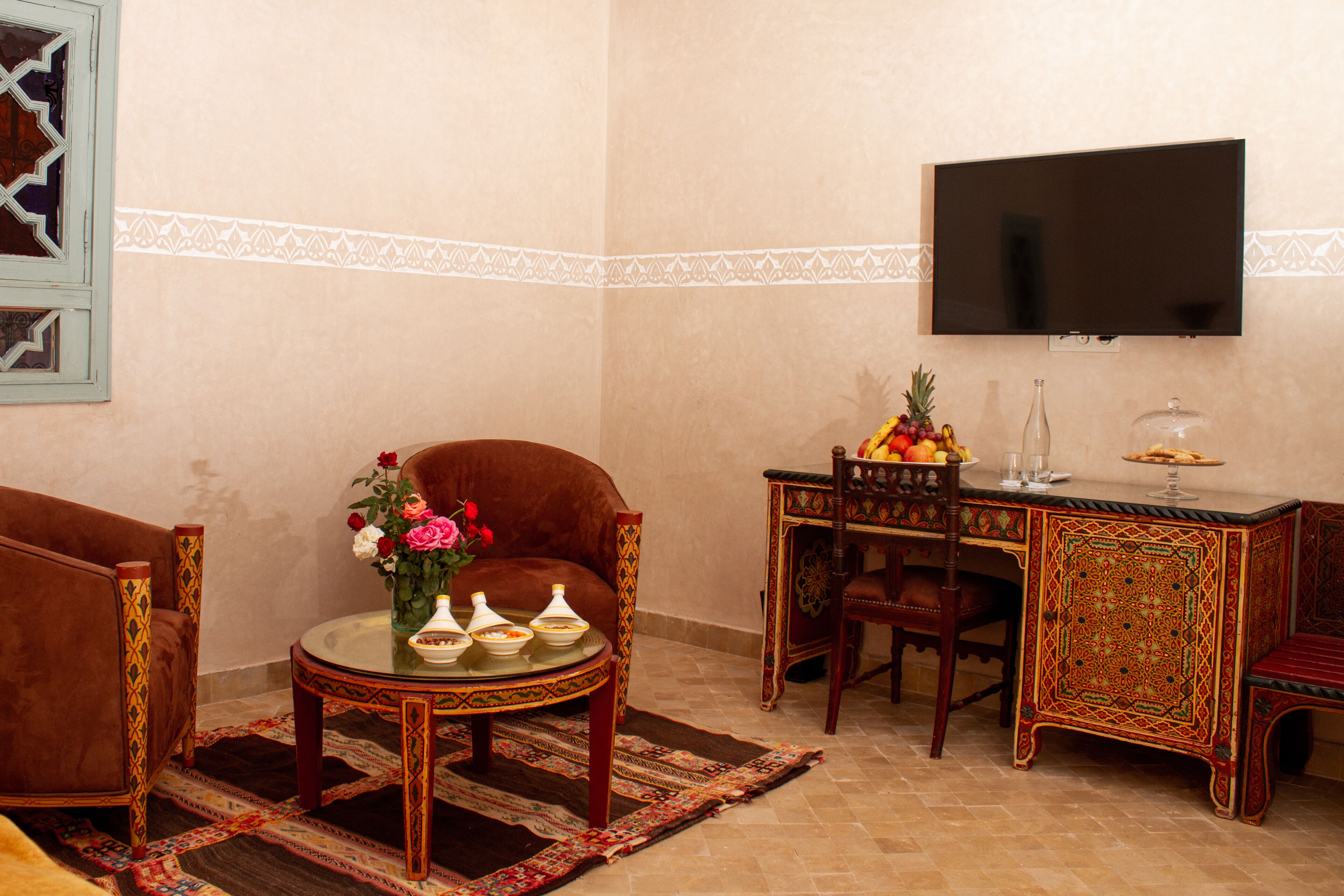 Ein Wohnzimmer mit einer Couch, einem Couchtisch mit Blumen, einem an der Wand montierten Fernseher und einem Dekorationstisch mit Obst und einem Glas.