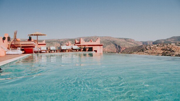 A clear blue pool with lounge chairs and umbrellas, surrounded by a red building with a view of hills. - Borj Des Cascades (Ait Taguella)