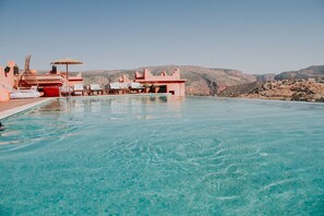 A clear blue pool with lounge chairs and umbrellas, surrounded by a red building with a view of hills. - Borj Des Cascades (Ait Taguella)