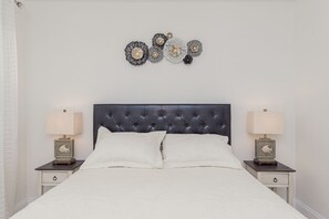 Un dormitorio con cabecera capitonada, dos mesitas de noche con lámparas, cama con ropa blanca y decoración en la pared.
