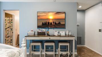 A room with a bed, a desk with a computer, a chair, and a wall-mounted TV displaying a sunset scene.