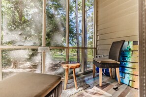 Una habitación con ventanas amplias, un taburete de madera, una silla y un mueble de colores.