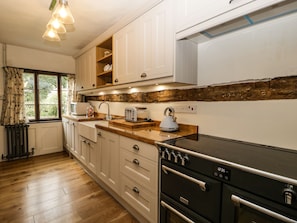 A modern kitchen with wooden cabinets, a black stove, and a window with curtains. - Churchend Cottage (Tewkesbury)
