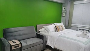 A room with a grey sofa, a small bedside table with a remote control, a bed with white bedding, and a green accent wall. - A Lovely Modern Retreat for Your Perfect Stay (Hayward)