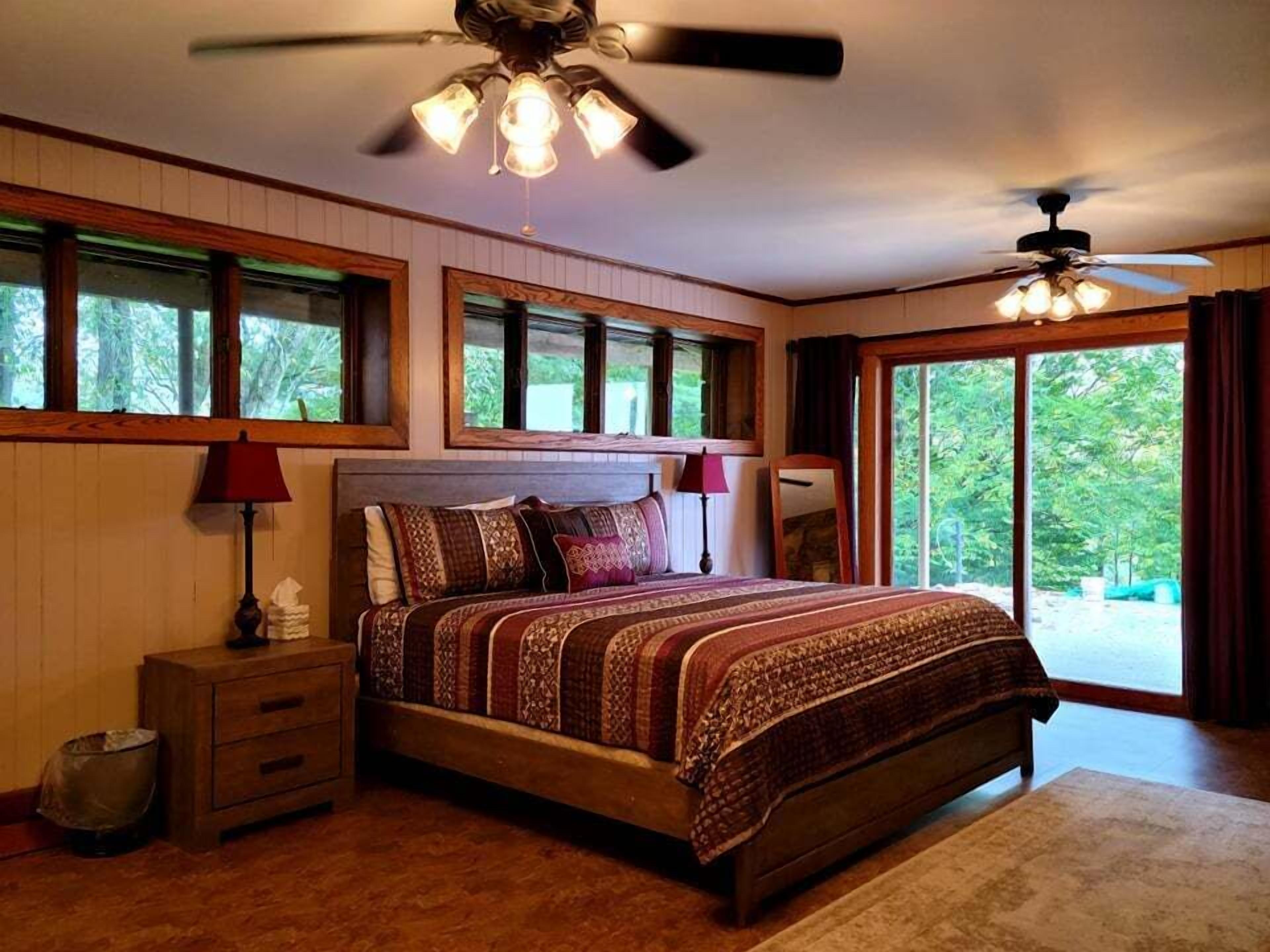 A bedroom with a large bed, two bedside lamps, a ceiling fan, and a view of greenery through the windows.