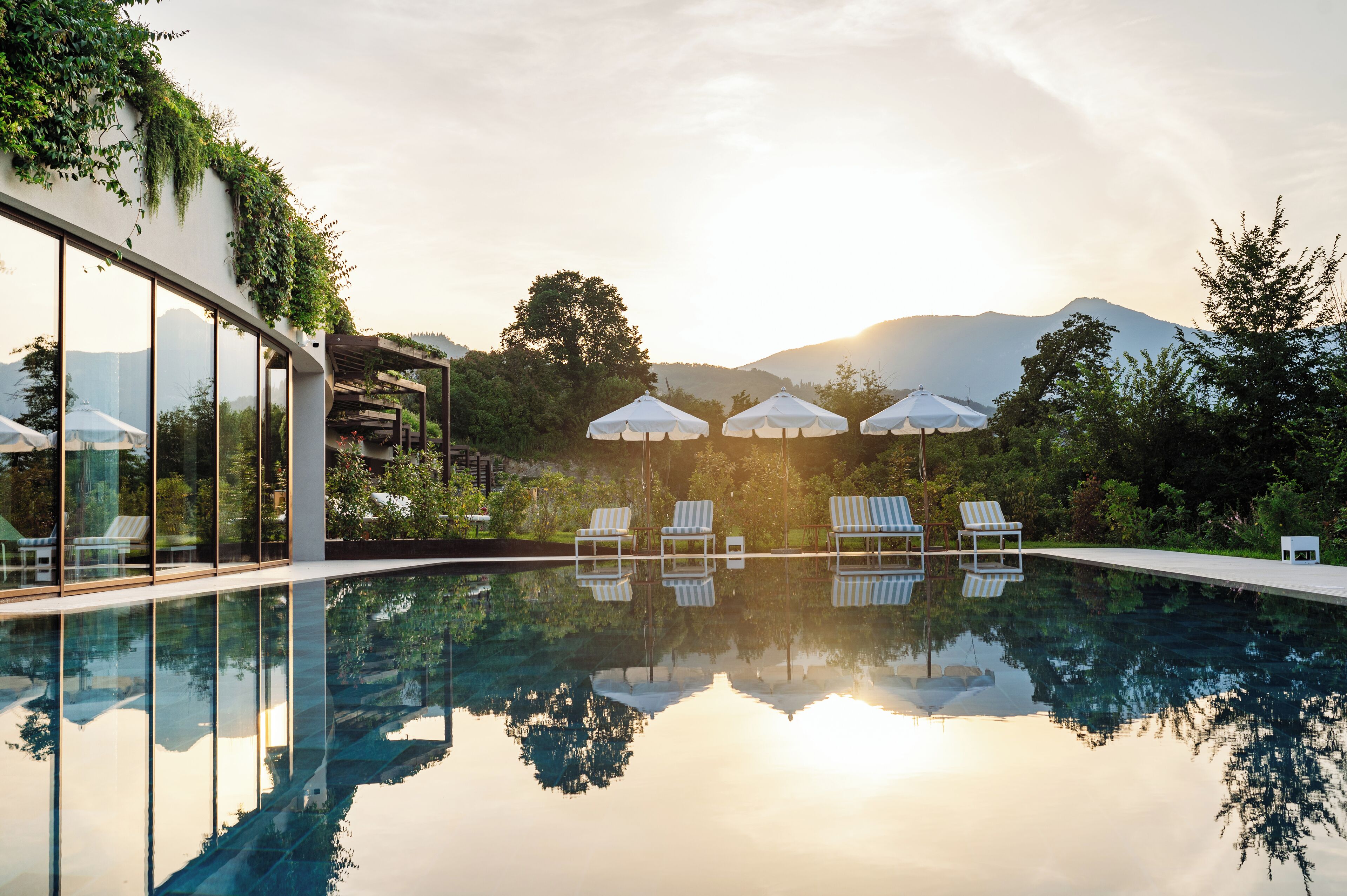Una piscina con sillones de descanso y sombrillas, rodeada de vegetación y montañas.